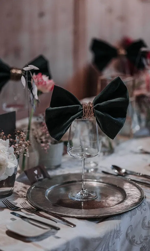 Detailaufnahme eines festlich gedeckten Platzes mit Weinglas, dekoriert mit einer schwarzen Stoffschleife und elegantem Metallteller.