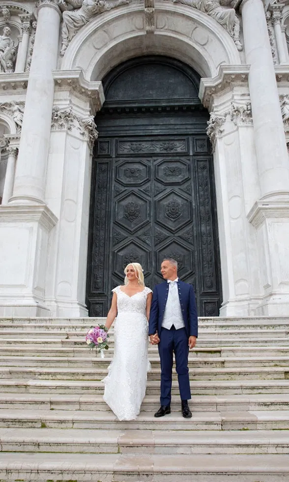 Braut und Bräutigam stehen Hand in Hand auf den Stufen vor einer prunkvollen venezianischen Kirche, die Braut im eleganten Spitzenbrautkleid mit Blumenstrauß, der Bräutigam im dunkelblauen Anzug – ein klassischer Hochzeitsmoment vor imposanter Architektur.