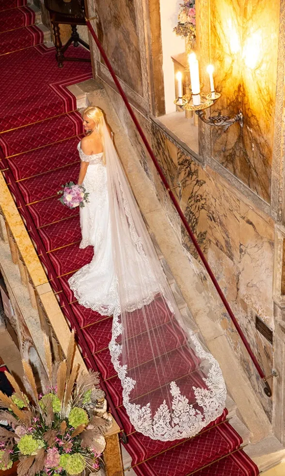 Braut steht auf einer mit rotem Teppich belegten Treppe im Inneren einer historischen Kirche, ihr Spitzenbrautkleid mit langem Schleier und ausladender Schleppe zieht sich elegant über die Stufen – ein atmosphärischer Hochzeitsmoment aus der Vogelperspektive.