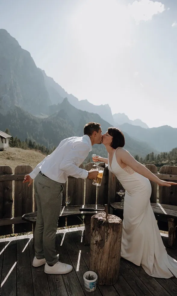 Braut und Bräutigam küssen sich auf einer Holzterrasse in den Bergen, während er ein Bierglas in der Hand hält.