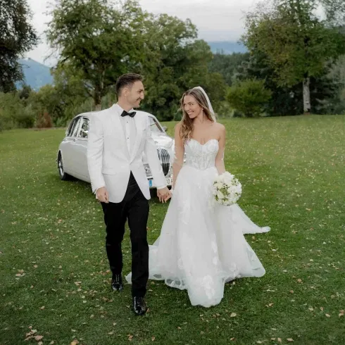 Brautpaar in eleganter Hochzeitskleidung geht Hand in Hand über eine grüne Wiese, im Hintergrund ein klassisches Hochzeitsauto und Naturkulisse.