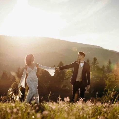 Brautpaar hält sich bei goldenem Abendlicht auf einer blühenden Wiese an den Händen, umgeben von Bergen und Natur – romantisches Hochzeitsshooting im Sonnenuntergang.