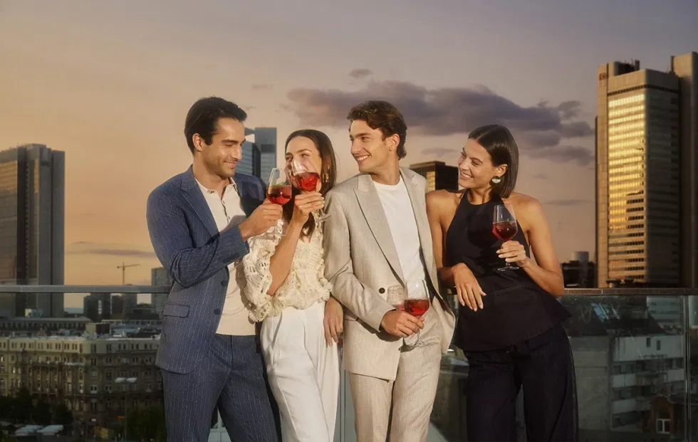 Freundesgruppe in Digel-Anzügen und modernen Outfits, die bei Sonnenuntergang auf einer Rooftop-Bar anstoßen; stilvolle Abendstimmung und elegante Menswear.