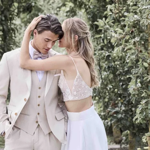 Braut und Bräutigam in modernem Hochzeitsoutfit umarmen sich in einer grünen Gartenkulisse.