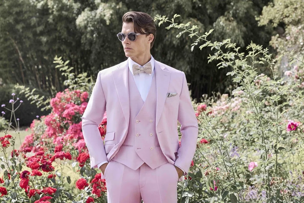 Bräutigam in rosafarbenem Hochzeitsanzug mit Fliege steht stilvoll in einem blühenden Garten.