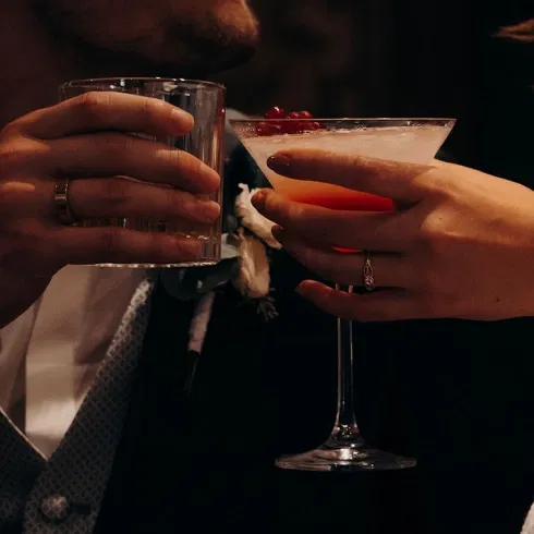 Braut und Bräutigam stoßen mit einem roten Cocktail und einem Whiskyglas bei stimmungsvoller Beleuchtung an