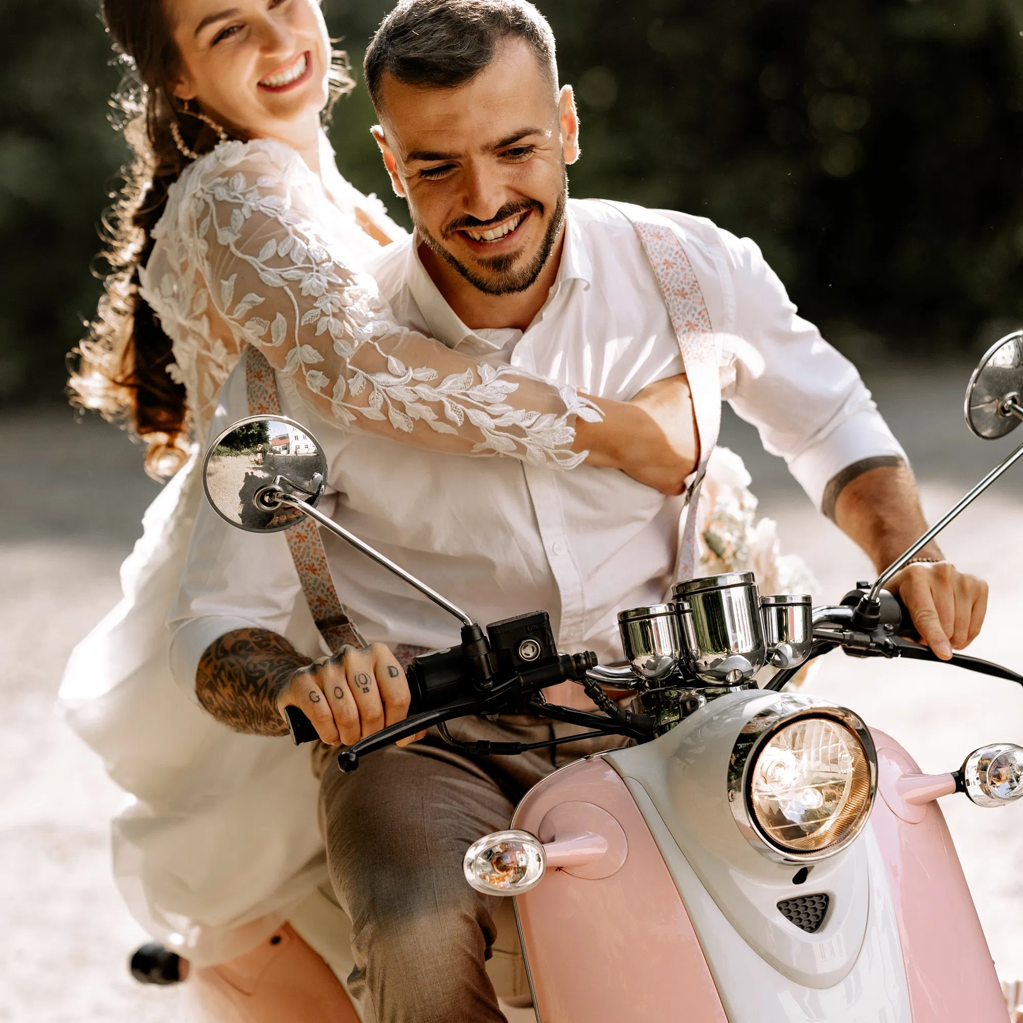 Brautpaar fährt lachend auf rosa Vespa; Braut im Spitzenkleid umarmt Bräutigam – Vespa als Hochzeitsfahrzeug.
