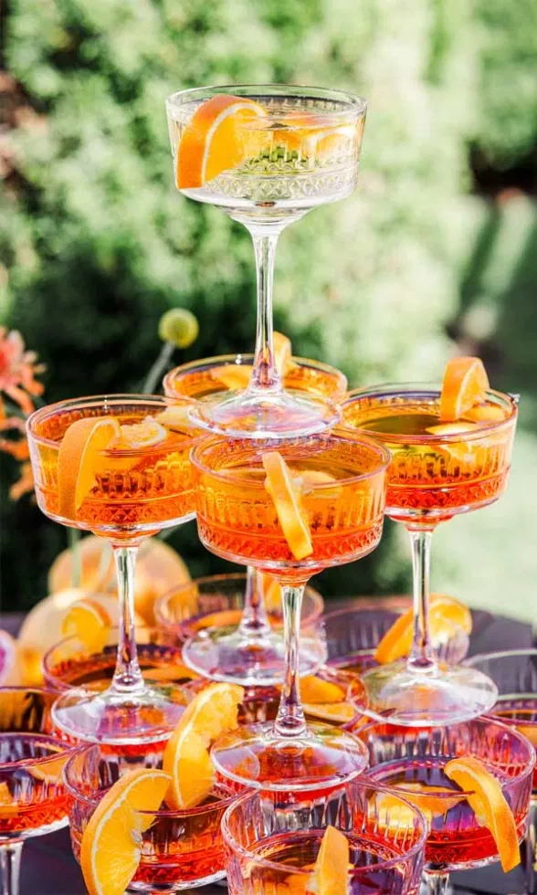 Gestapelte Coupette-Gläser mit leuchtend orangefarbenem Aperitif und frischen Orangenscheiben als Garnitur, arrangiert als stilvoller Drink-Tower vor grünem, sommerlichem Hintergrund.