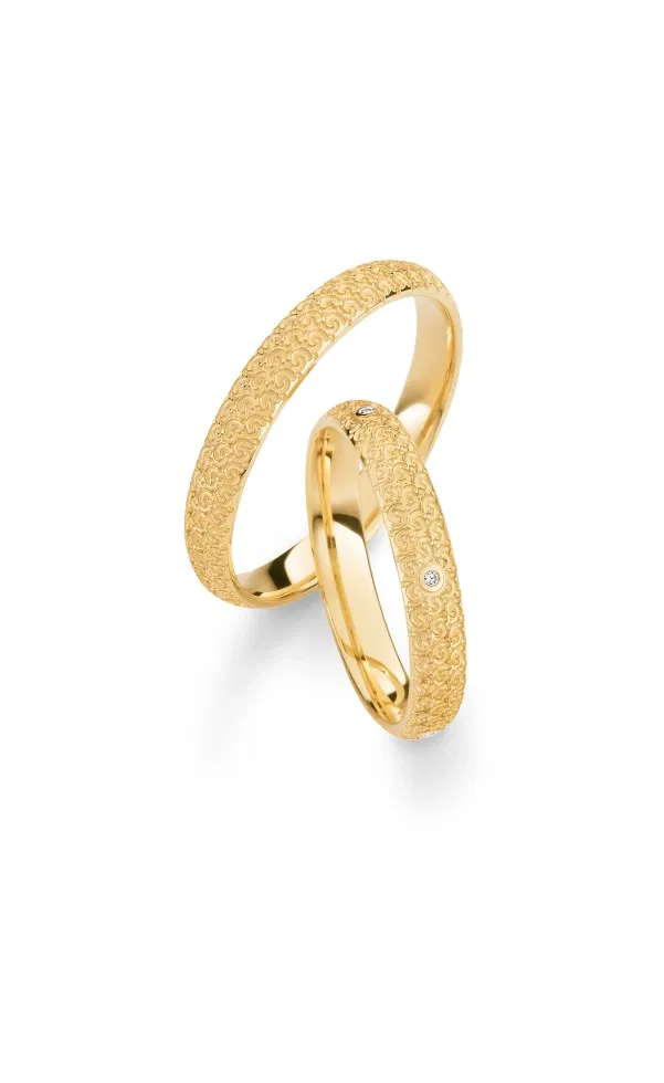 Zwei Eheringe aus Gelbgold mit feiner, ornamentaler Struktur im Vintage-Stil, einer davon mit dezent gefasstem Diamanten, elegant vor hellem Hintergrund fotografiert.
