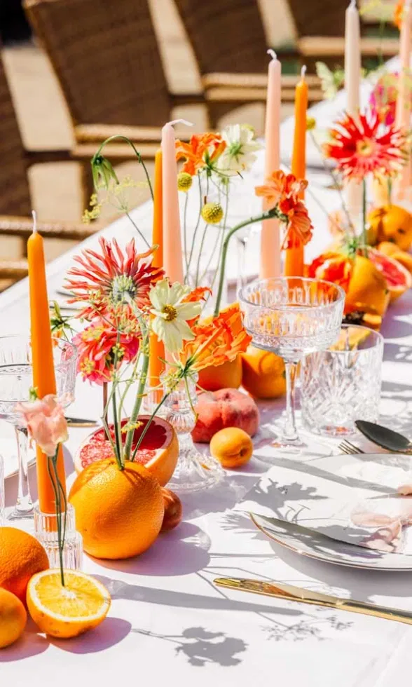 Lange, festlich gedeckte Hochzeitstafel mit orangefarbenen Kerzen, leuchtenden Sommerblumen, frischen Zitrusfrüchten und elegantem Glas- sowie Goldbesteck in sonniger Outdoor-Atmosphäre.