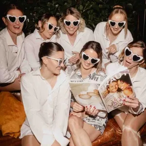 Fröhliche Brautjungfern in weißen Blusen tragen herzförmige Sonnenbrillen und sitzen gemeinsam auf einem Sofa, während sie lachend ein Hochzeitsmagazin betrachten – ausgelassene Getting-Ready-Stimmung vor der Hochzeit.
