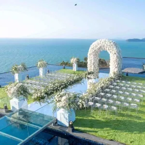Elegante Outdoor-Trauung am Meer mit weißem Blumenbogen, floraler Dekoration und transparenten Stühlen auf einer Rasenfläche mit Poolblick – luxuriöse Destination Wedding mit Panorama-Ozeanblick.