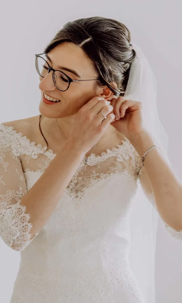 Braut mit Brille steckt sich lächelnd den Ohrring an, trägt ein Spitzenbrautkleid mit Schleier, aufgenommen vor hellem, ruhigem Hintergrund.
