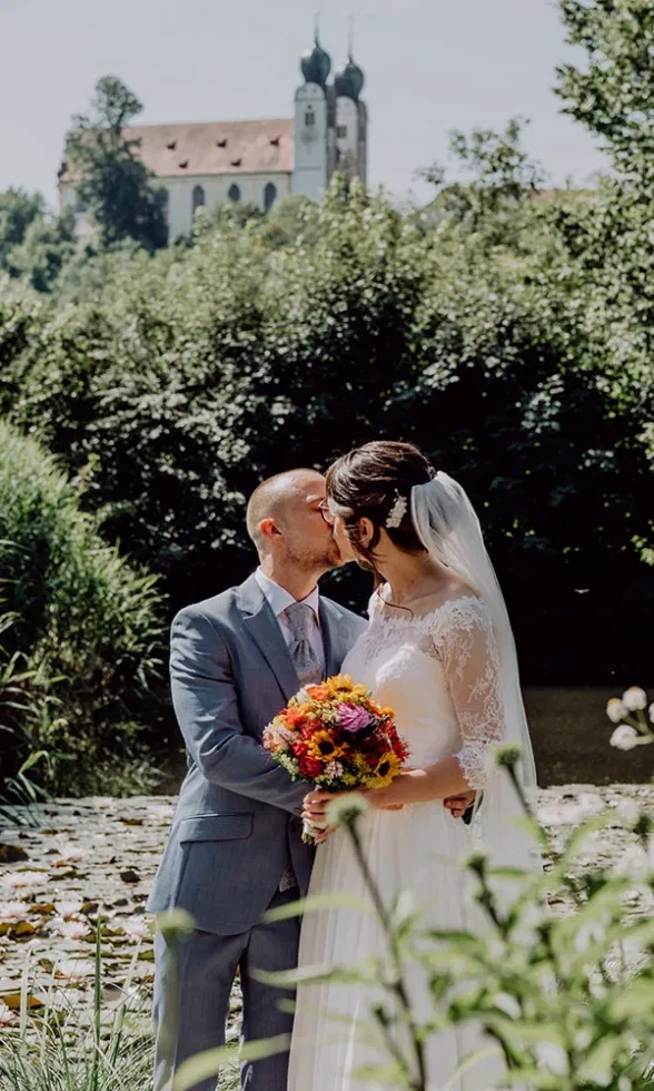 Brautpaar küsst sich am Wasser, im Hintergrund eine Kirche, die Braut hält einen farbenfrohen Blumenstrauß und trägt ein Spitzenbrautkleid mit Schleier.