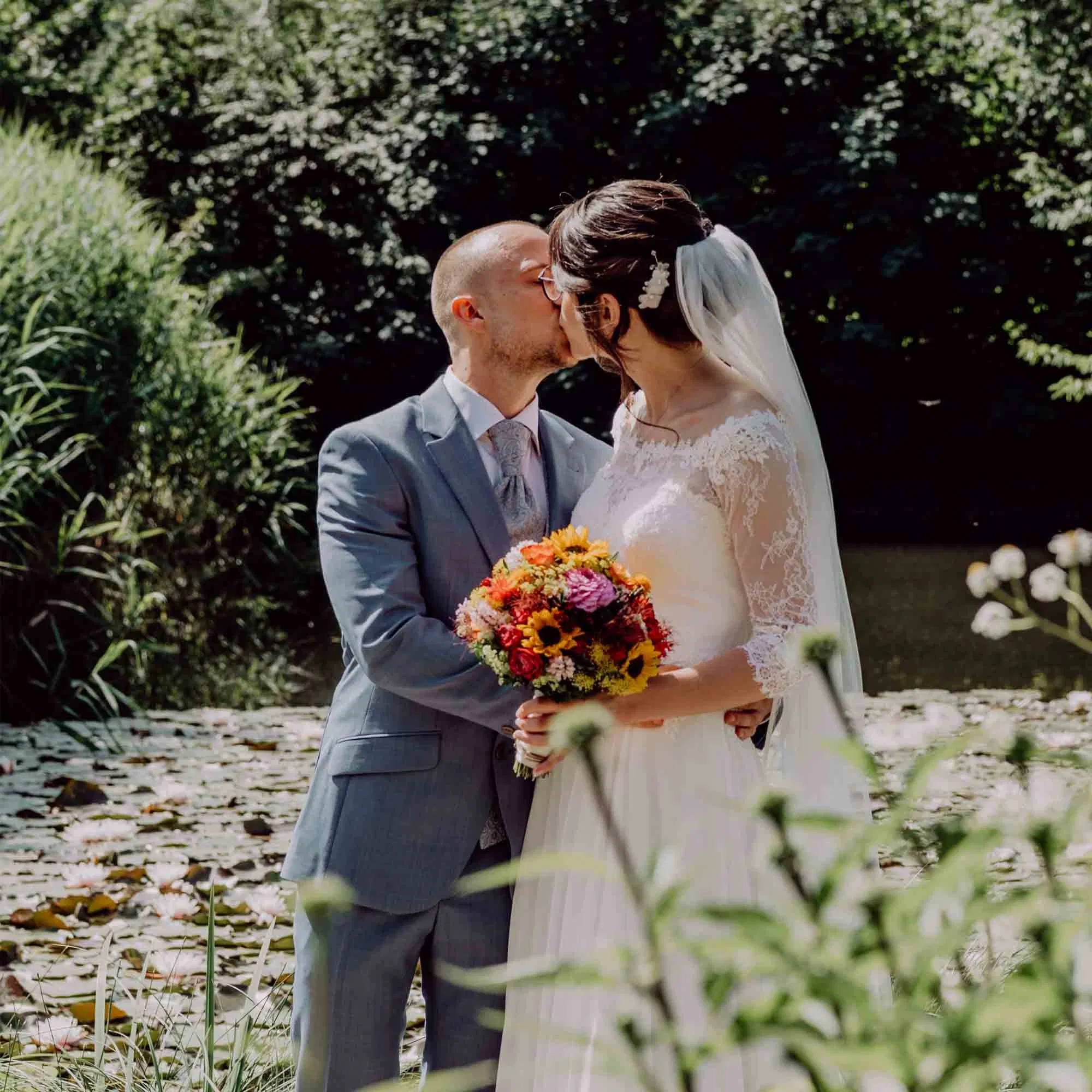 Brautpaar küsst sich im Grünen, die Braut trägt ein langärmeliges Spitzenbrautkleid mit Schleier und hält einen farbenfrohen Herbststrauß, der Bräutigam einen hellblauen Anzug.
