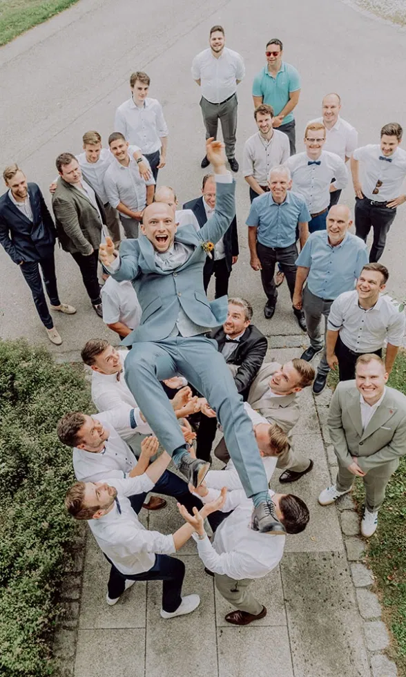 Ausgelassener Hochzeitsmoment: Der Bräutigam wird von seinen Freunden lachend in die Luft geworfen, umgeben von der Hochzeitsgesellschaft im Freien.