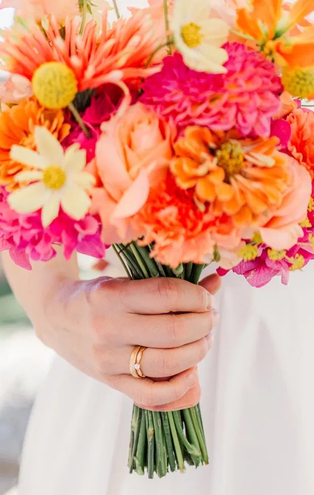 Braut hält farbenfrohen Strauß in Orange, Pink und Gelb – sommerliche Blumen für die Gartenhochzeit