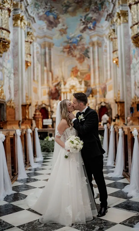 Brautpaar küsst sich im Mittelgang einer prunkvollen Kirche, die Braut trägt ein fließendes Brautkleid mit floralen Applikationen, umgeben von kunstvollen Fresken und warmem Licht.