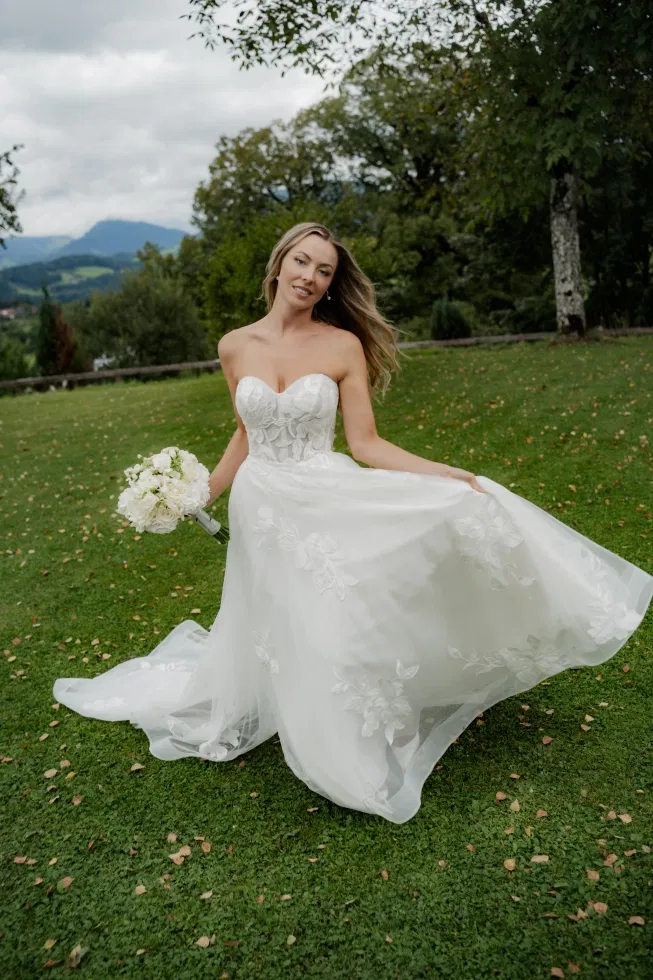 Braut in trägerlosem, floralen Brautkleid steht auf einer grünen Wiese und hält einen weißen Brautstrauß, während sie den Rock ihres Kleides elegant in Bewegung bringt, umgeben von Natur und sanfter Bergkulisse im Hintergrund.