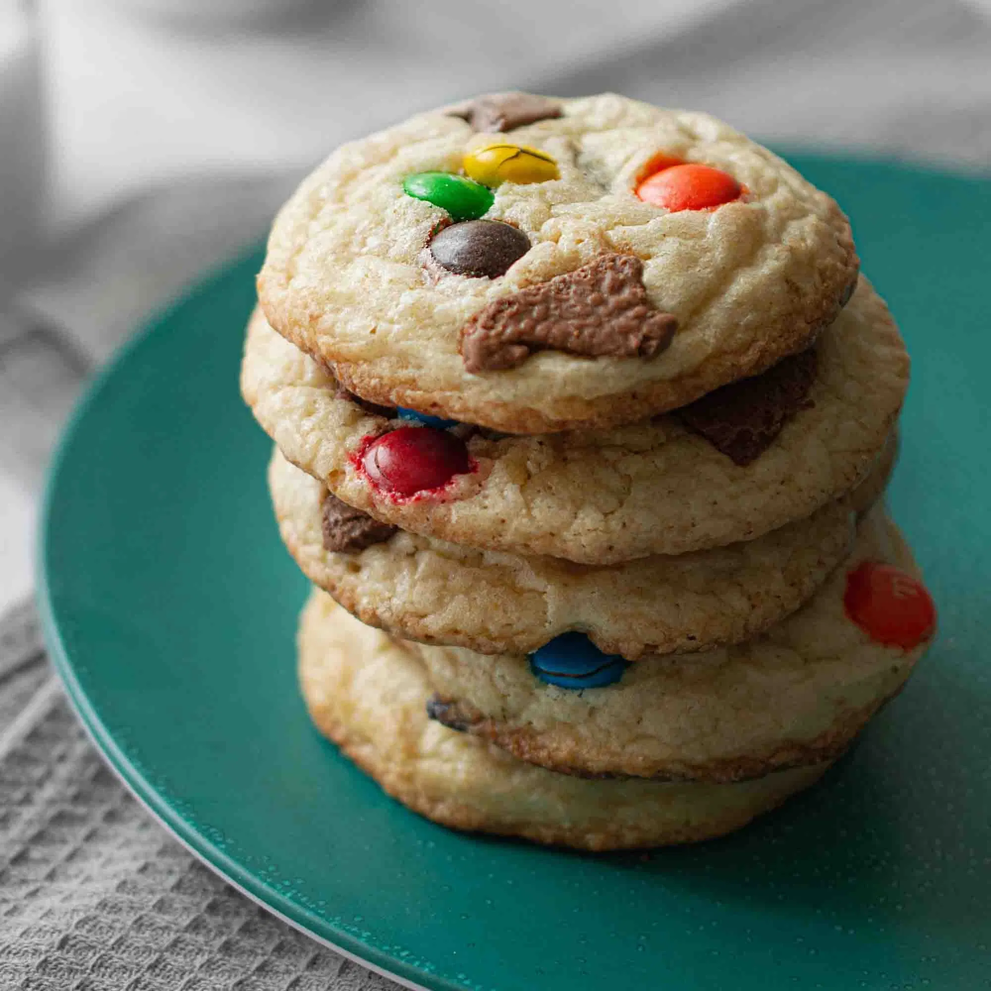 Gestapelte Cookies mit bunten Schokolinsen und Schokoladenstücken auf einem grünen Teller.