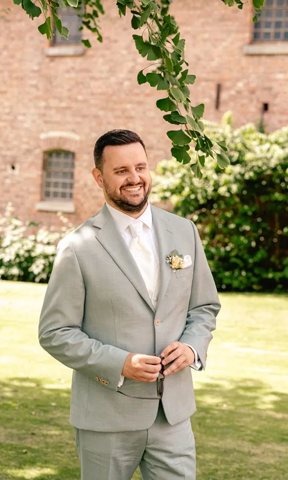 Bräutigam im hellgrauen Anzug mit weißer Krawatte und Anstecker, lächelnd im Grünen vor einer Backsteinfassade während der Hochzeitsfeier.
