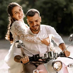 Braut und Bräutigam fahren lachend gemeinsam auf einem Roller, die Braut im Spitzenkleid umarmt den Bräutigam von hinten – lockeres Hochzeitsfoto mit Vintage-Flair und sommerlicher Leichtigkeit.