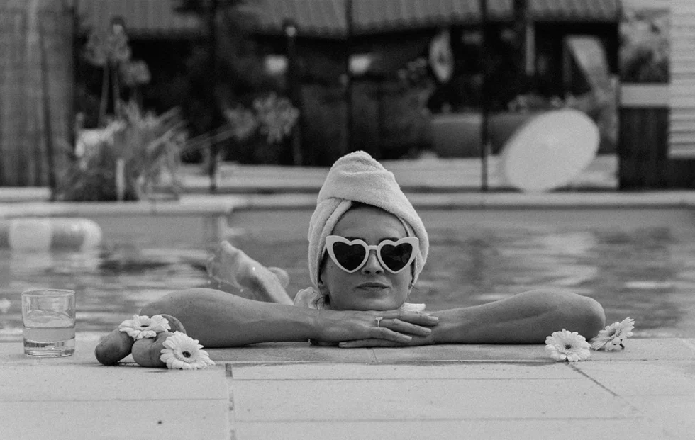 Schwarzweißaufnahme einer Braut im Pool mit Sonnenbrille, Blumen und Drink – ruhiger Moment vor dem großen Hochzeitstag.