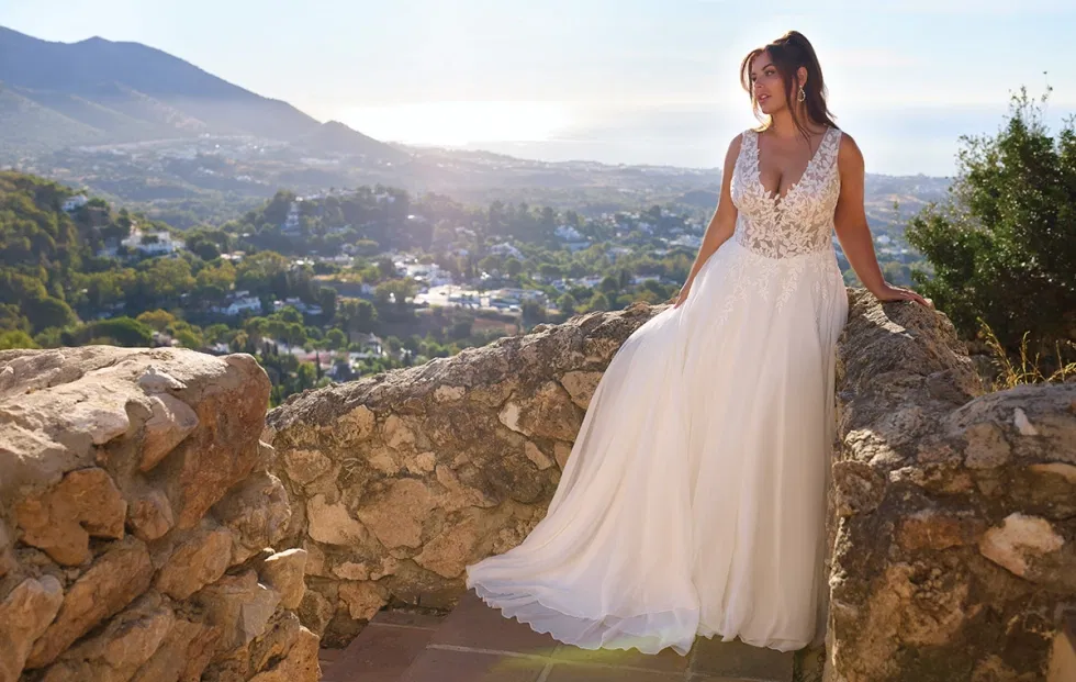 Braut im fließenden A-Linien-Brautkleid mit Spitzenoberteil, inszeniert auf einer Natursteinmauer mit mediterraner Landschaft im Hintergrund.