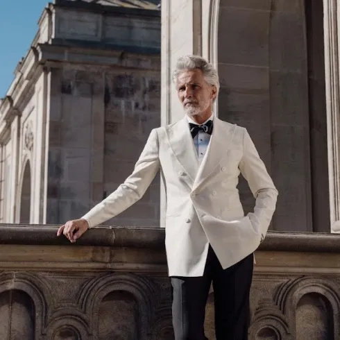 Eleganter Bräutigam im weißen Smoking mit schwarzer Fliege vor historischer Architektur – stilvolles Hochzeitsmode-Shooting bei Sonnenschein.