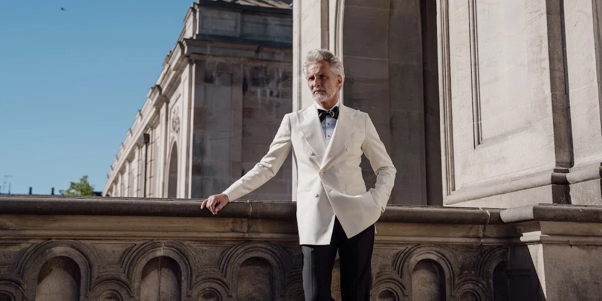 Eleganter Bräutigam im weißen Smoking mit schwarzer Fliege vor historischer Architektur – stilvolles Hochzeitsmode-Shooting bei Sonnenschein.