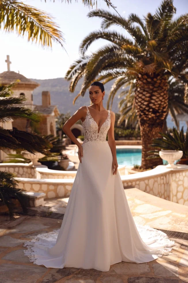 Elegantes A-Linien-Brautkleid mit tiefem V-Ausschnitt und floraler Spitzenapplikation, präsentiert in einer luxuriösen Villa mit Pool und Palmen.