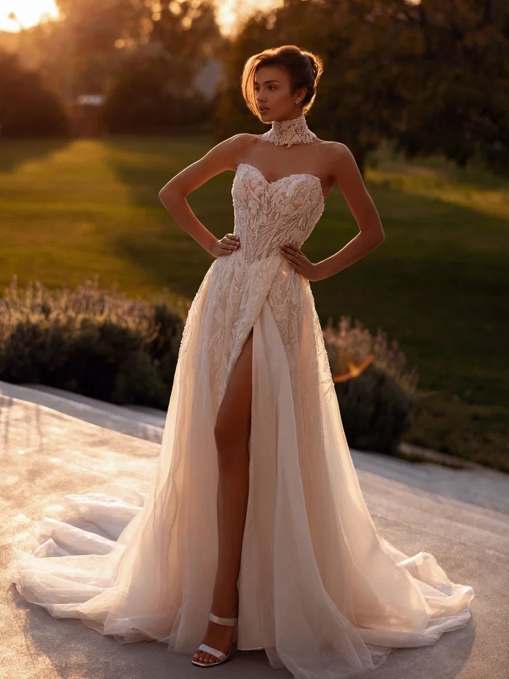 Luftiges A-Linien-Brautkleid mit floraler Spitze, langen transparenten Ärmeln und hohem Beinschlitz, aufgenommen auf einer Terrasse mit Blick auf das Meer.