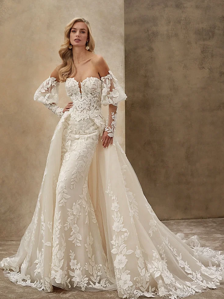Romantisches Off-Shoulder-Brautkleid in Fit-and-Flare-Silhouette mit floraler 3D-Spitze, transparenten Ärmeln und abnehmbarer Tüllschleppe vor neutralem Studiohintergrund.