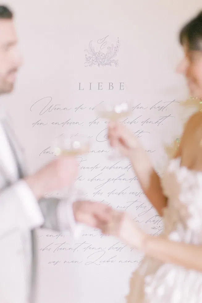 Brautpaar stößt mit Champagnergläsern an, im Hintergrund ein handgeschriebener Text über Liebe in elegantem Fine-Art-Stil.