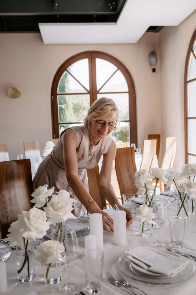 Wedding-Planerin dekoriert einen festlich gedeckten Hochzeitstisch mit weißen Blumen, Kerzen und eleganter Papeterie in einer hellen Hochzeitslocation.