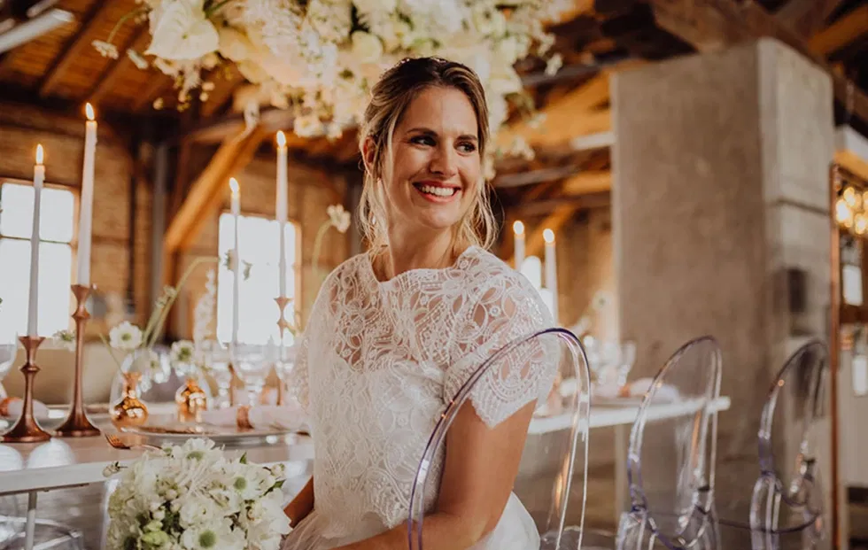 Lachende Braut im Spitzenkleid sitzt an einer langen Hochzeitstafel im Sägewerk am Mondsee, warmes Licht, Kupferdetails und transparente Stühle schaffen eine moderne Hochzeitsatmosphäre.