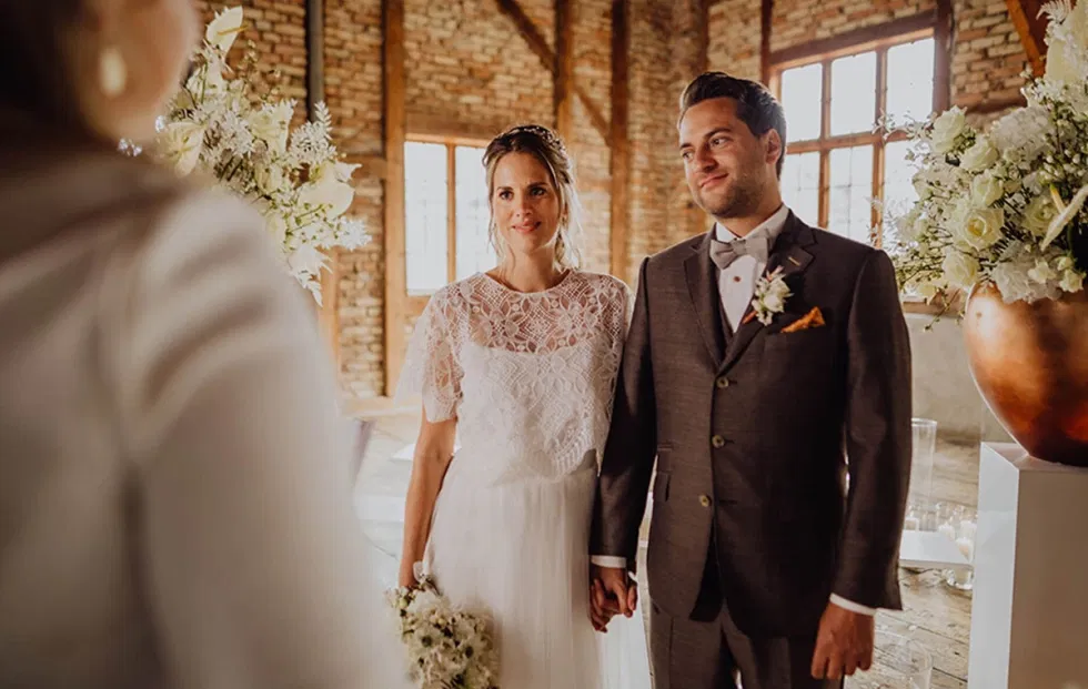 Braut und Bräutigam stehen Hand in Hand während einer freien Trauung im Sägewerk am Mondsee, eingerahmt von floralen Arrangements und historischem Industrieambiente.