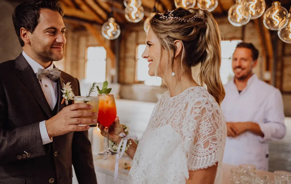 Braut und Bräutigam stoßen mit Cocktails in der Hochzeitslocation Sägewerk am Mondsee an, moderne Lichtinstallationen und offenes Hallenkonzept im Hintergrund.