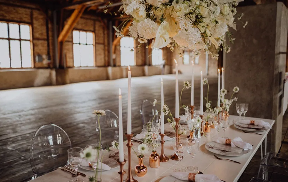 Detailaufnahme einer festlich gedeckten Hochzeitstafel im Sägewerk am Mondsee mit Kerzen, Glasvasen, Blumen und Blick in die weitläufige Eventhalle.