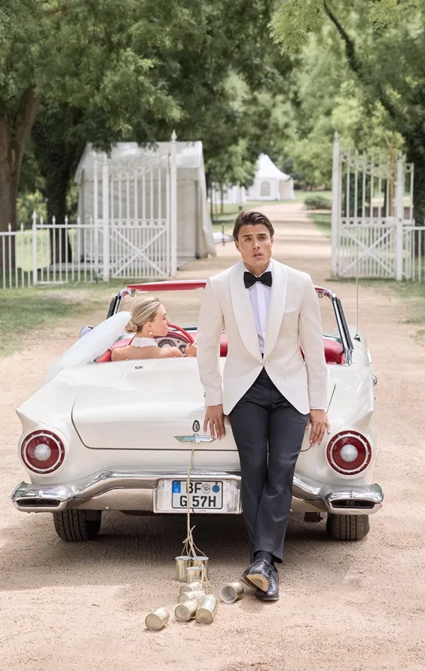 Bräutigam im weißen Smoking mit schwarzer Fliege vor einem Oldtimer – stilvoller Hochzeitslook mit Hollywood-Glamour.