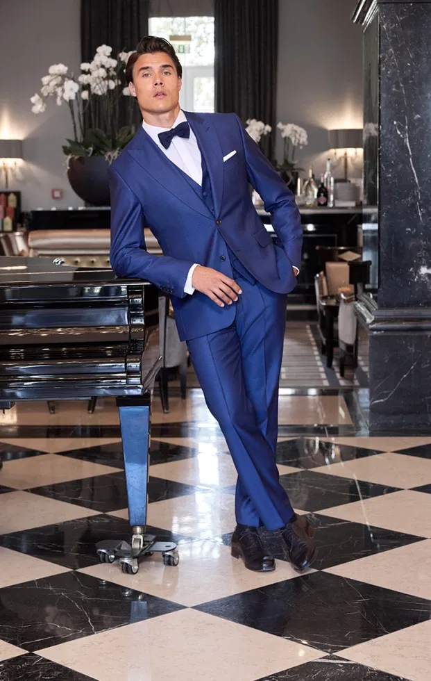 Bräutigam in royalblauem Anzug mit Fliege, elegant angelehnt an einen schwarzen Flügel in stilvollem Interieur.