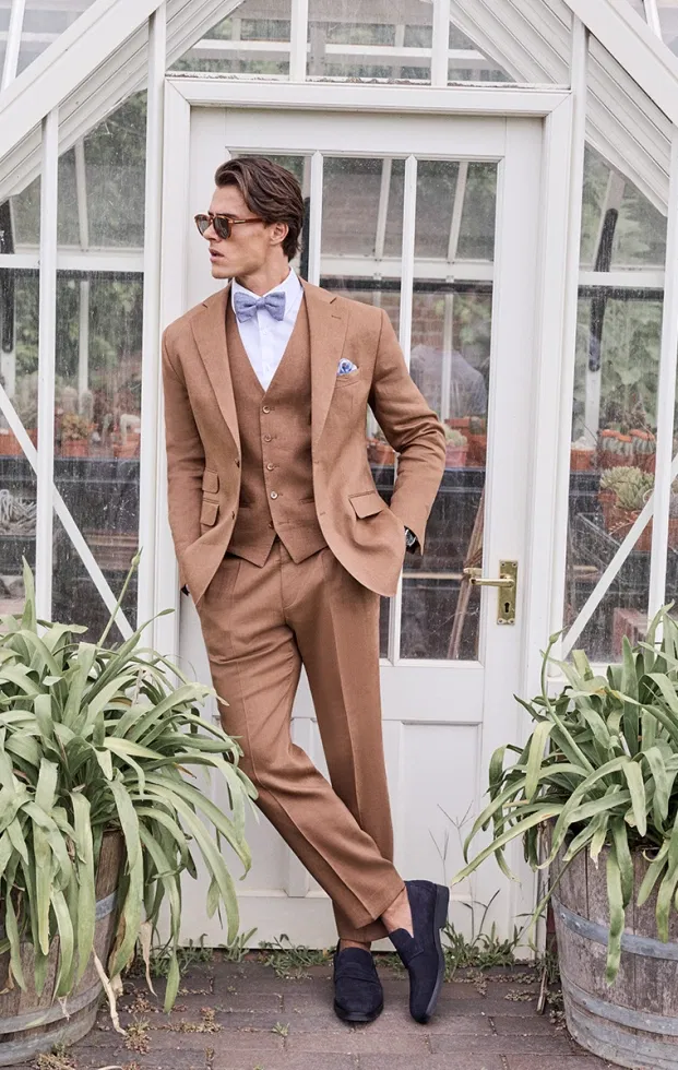 Kupferfarbener Dreiteiler mit blauer Fliege und Einstecktuch – eleganter Hochzeitsanzug mit Retro-Charme im Gewächshaus-Ambiente.