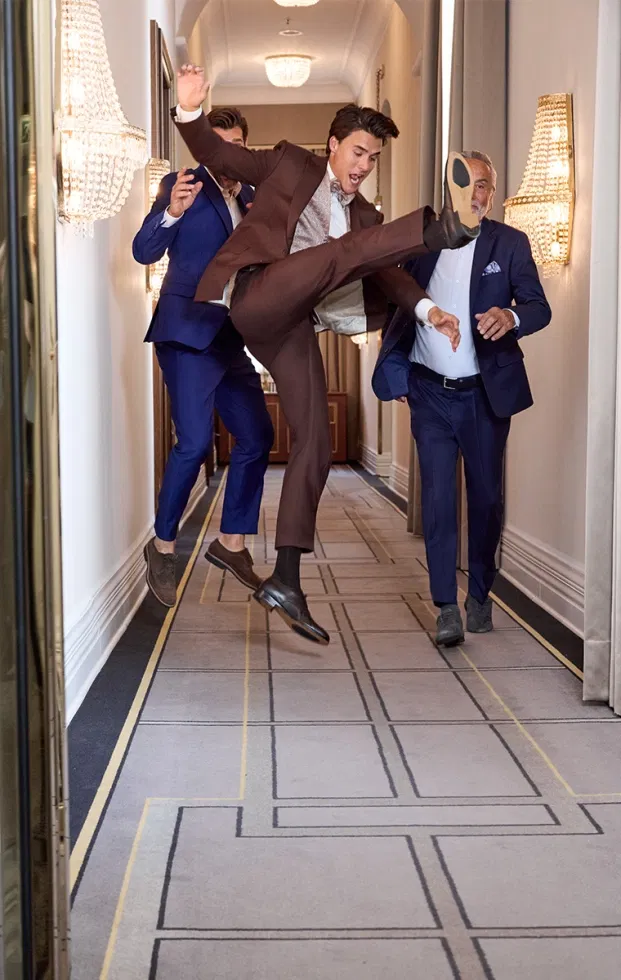 Bräutigam in braunem Anzug springt ausgelassen mit Freunden im Hotelgang – moderner Hochzeitsmoment voller Lebensfreude.