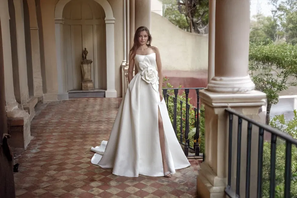 Elegantes Brautkleid aus Satin mit großer Blumenapplikation und Beinschlitz, Braut posiert in historischer Arkadengalerie – moderne Hochzeitsmode im mediterranen Stil.