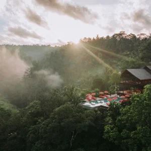 Luxuriöses Resort mit Infinity-Pool und roten Sonnenschirmen, eingebettet in eine üppige tropische Landschaft bei Sonnenaufgang mit Nebelschwaden über dem Dschungel.