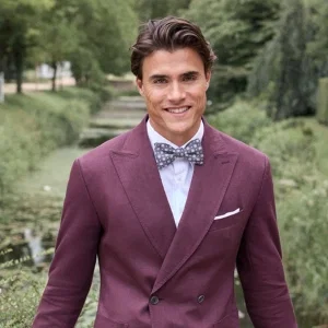 Ein lächelnder Mann in einem bordeauxfarbenen Anzug mit Fliege steht in einer grünen Parklandschaft an einem schmalen Wasserlauf.