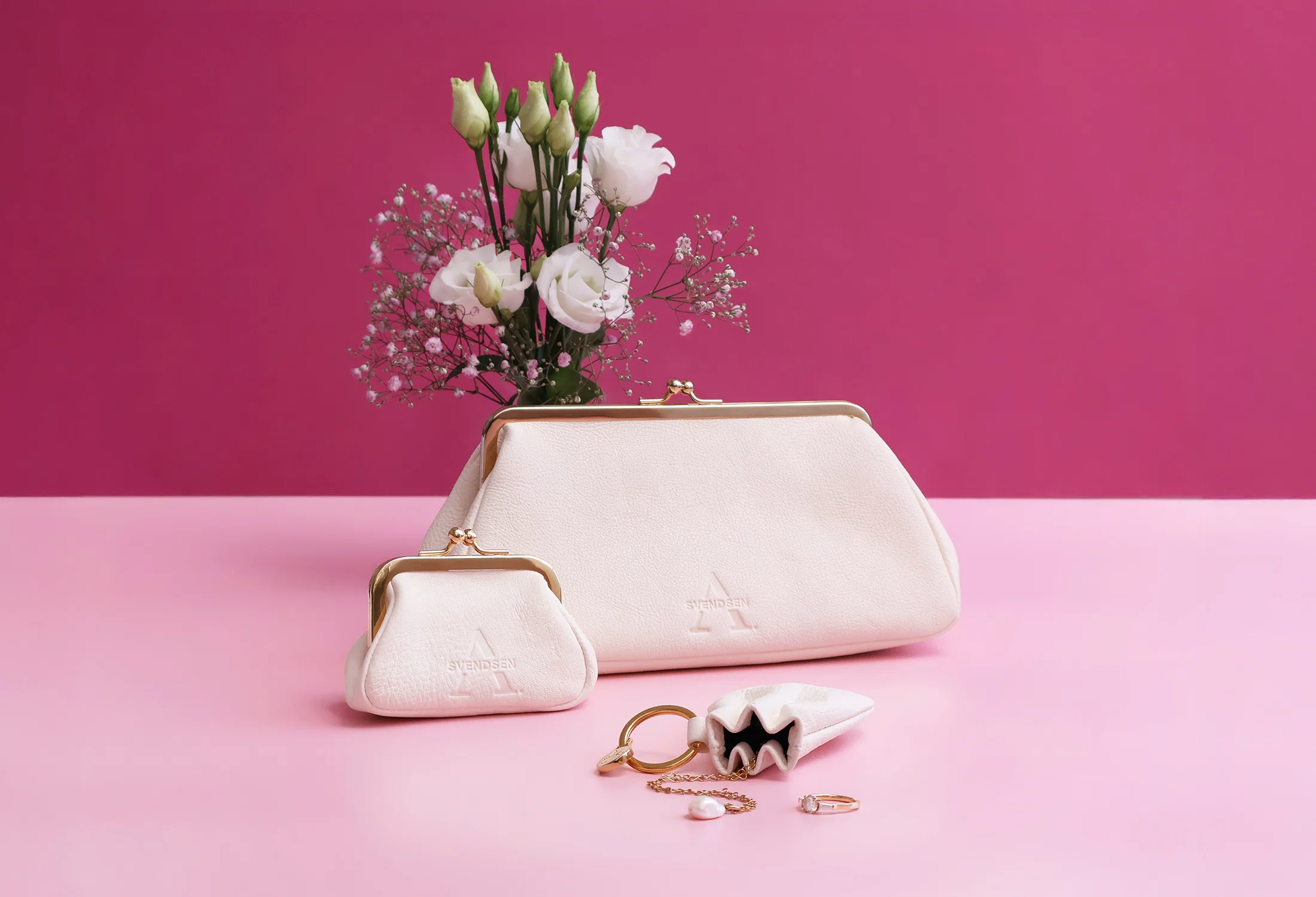 Elegantes Taschen-Set von Alexandra Svendsen mit weißer Abendtasche, kleinem Schmucktäschchen und Leder-Accessoire vor pinkem Hintergrund, dekoriert mit weißen Blumen und Schmuck.