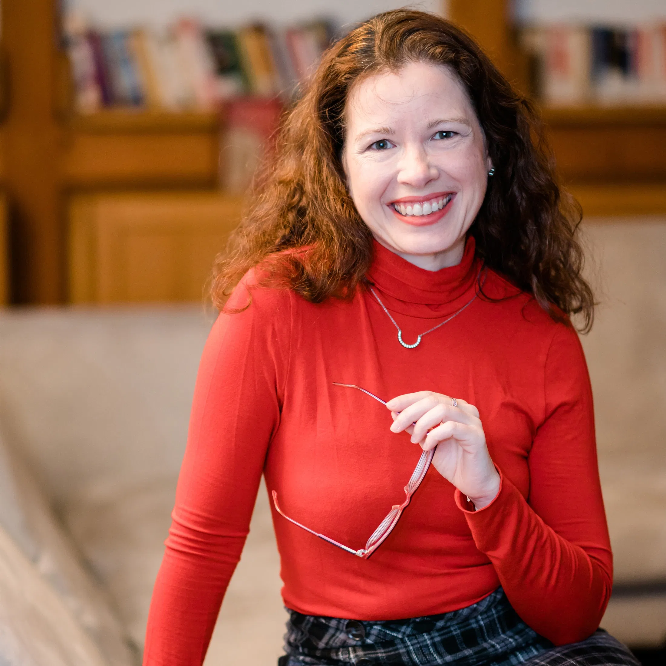 Porträt einer lächelnden Frau mit lockigem Haar in rotem Rollkragenpullover, die eine Brille in der Hand hält, vor einem Bücherregal im Hintergrund.