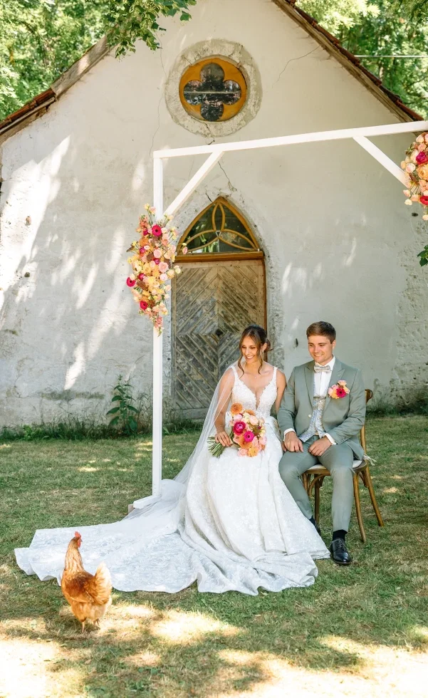 Brautpaar sitzt bei einer freien Trauung vor einer kleinen Kapelle, umrahmt von einem floralen Traubogen, während ein Huhn über die Wiese läuft.