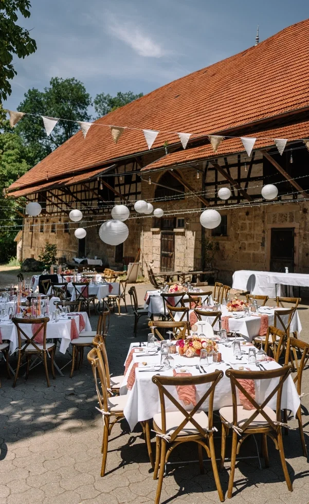 Rustikale Hochzeitslocation im Freien mit gedeckten Tischen, Holzstühlen und dekorativen Lichterketten vor einer Scheune.